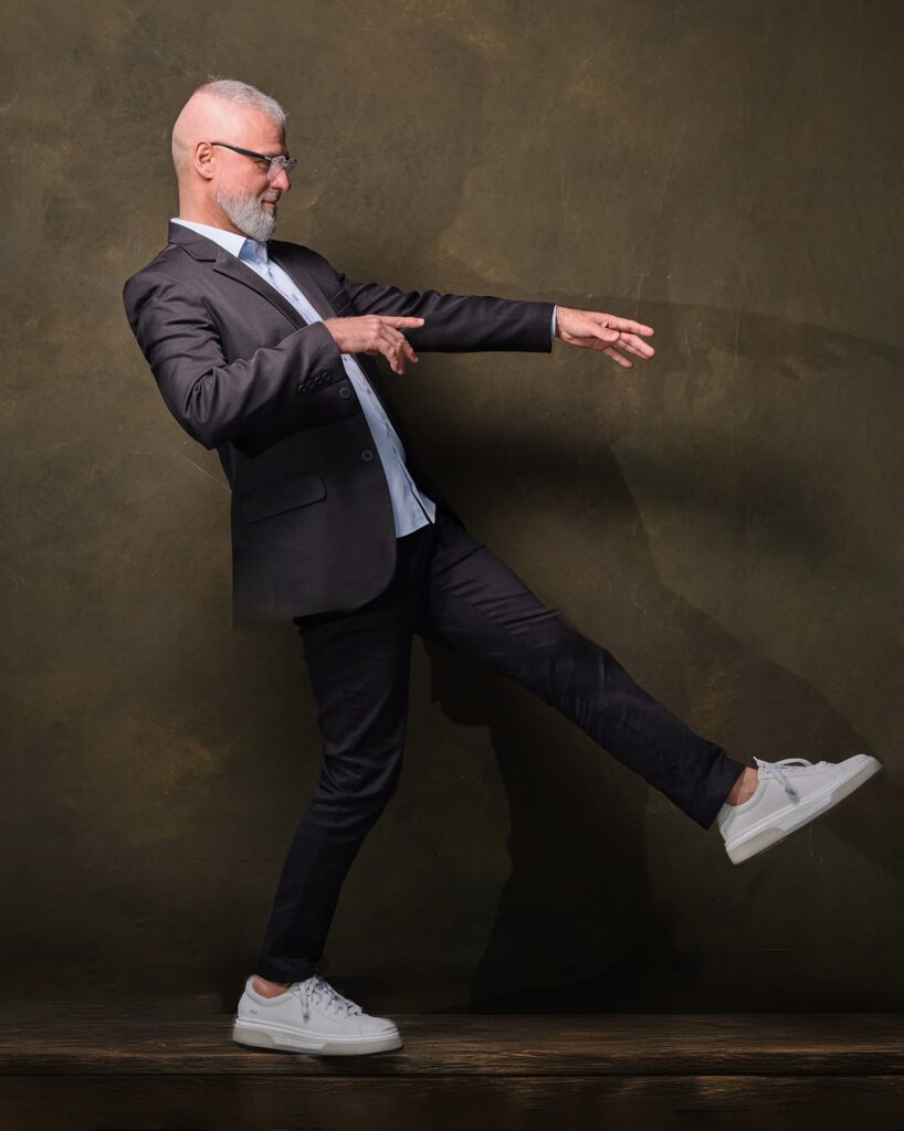 Homem de cabelos grisalhos e barba curta, usando óculos, blazer escuro, camisa clara, calça preta e tênis brancos. Ele está em pé diante de um fundo marrom texturizado, inclinado levemente para trás, com um braço estendido à frente e uma perna levantada, como se estivesse equilibrando-se ou simulando um movimento de dança ou gesto performático.