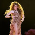 Pessoa em cena: uma cantora/dançarina em pleno movimento, com o corpo levemente inclinado e o gesto direcionado ao próprio tronco, sugerindo controle rítmico e consciência corporal típica de performances coreografadas. Expressão facial: forte e dramática, com a boca aberta e o olhar concentrado, transmitindo intensidade emocional e presença cênica. Figurino: vestido assimétrico em tons terrosos/dourados, com textura orgânica e aparência artesanal. A fenda lateral revela a perna e reforça a dinâmica do movimento, dialogando com dança e ritmo. Cabelo e movimento: cabelos longos e volumosos soltos, capturados em pleno deslocamento, ampliando visualmente a sensação de força e fluidez. Iluminação: luz quente, dourada, incidindo de cima ou por trás, criando um halo luminoso que destaca o corpo contra um fundo escuro, típico de grandes shows. Atmosfera geral: sensualidade, vigor e potência rítmica. A cena remete a uma estética que valoriza o corpo como instrumento musical e expressivo, onde dança e canto se fundem.